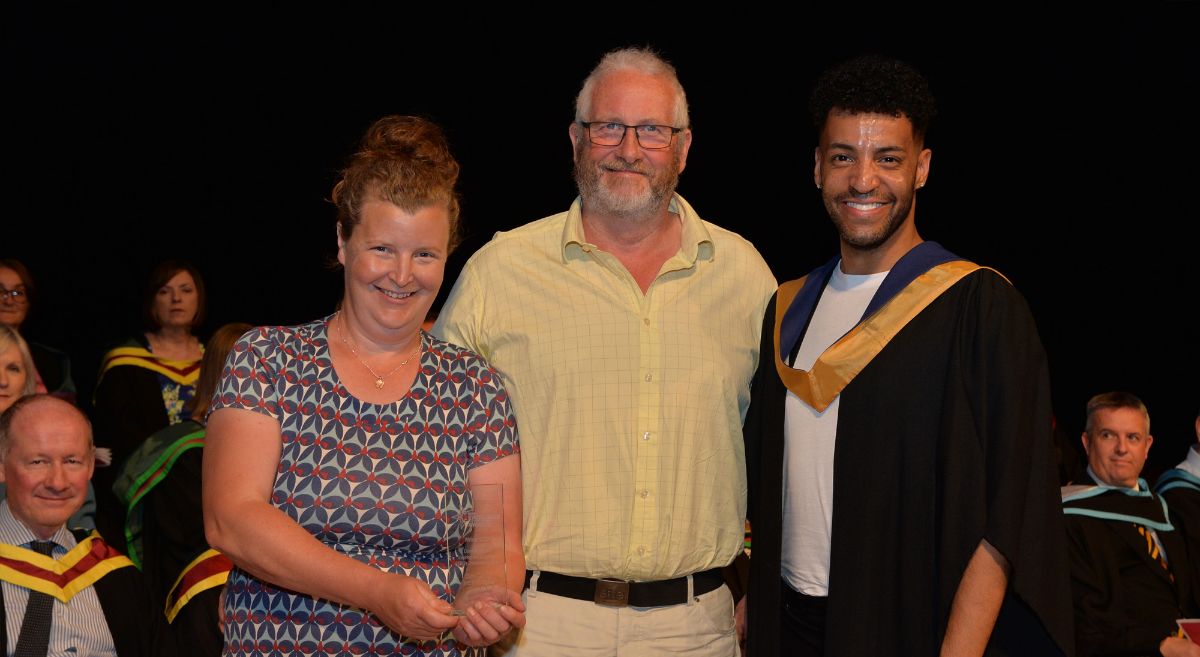 A woman in a pattered dress holding an award beside a man in a yellow shirt and a man in graduation gown